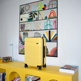 A yellow, hard-surface suitcase in light yellow sits on an ochre bookcase with fanciful shapes, right under a hand-sprayed canvas.