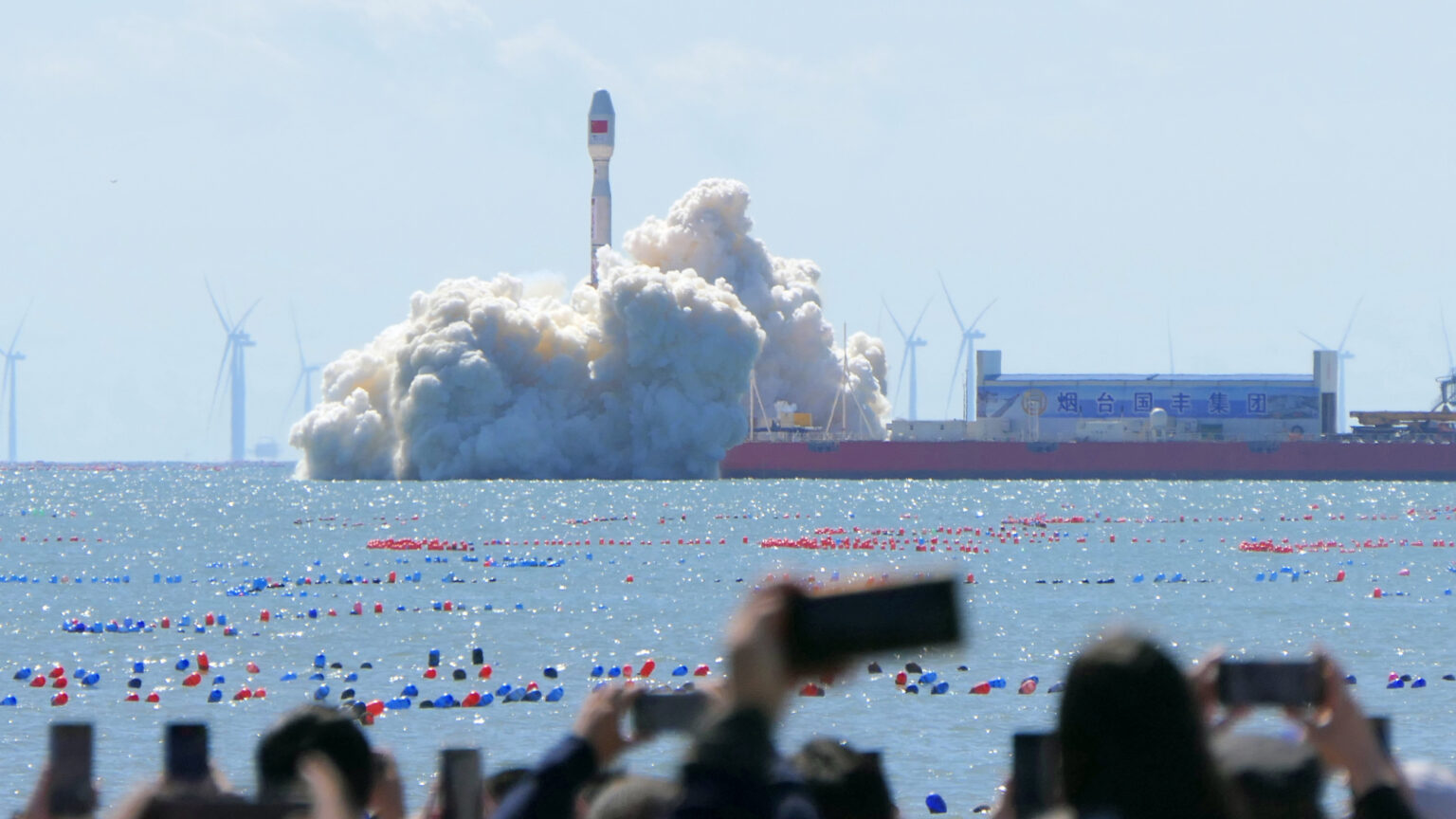 spectators watch a white rocket launch into a blue sky from a ship anchored at sea, not far offshore