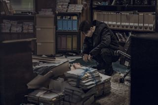 A man (Park Seo-jun as Jang Ho-jae) lies on the floor of a crowded office, partly blocked by piles of newspapers, as another man (Bae Hyeon-seong as Seung-jo) crouches over him, in Gyeongseong Creature S2.