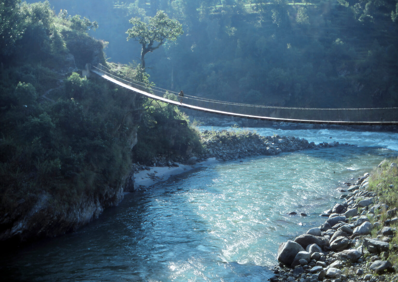 a river with a suspension bridge over it with a man walking across, with trees surrounding the gorge