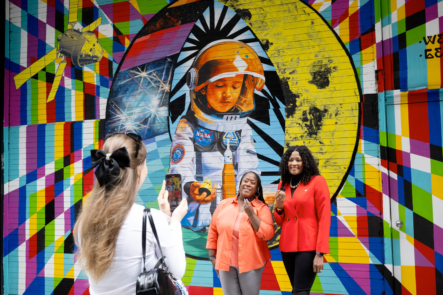 two women stand in front of a colorful space exploration mural while a third woman records video of them on her phone