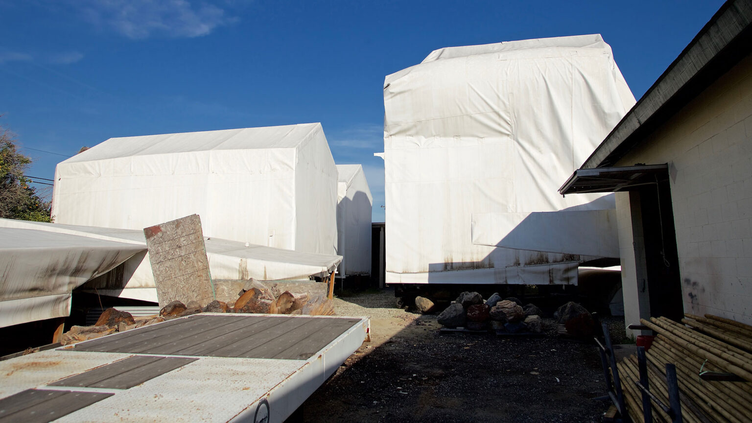 three large pieces of equipment covered by white tarps sit in a sunny lot