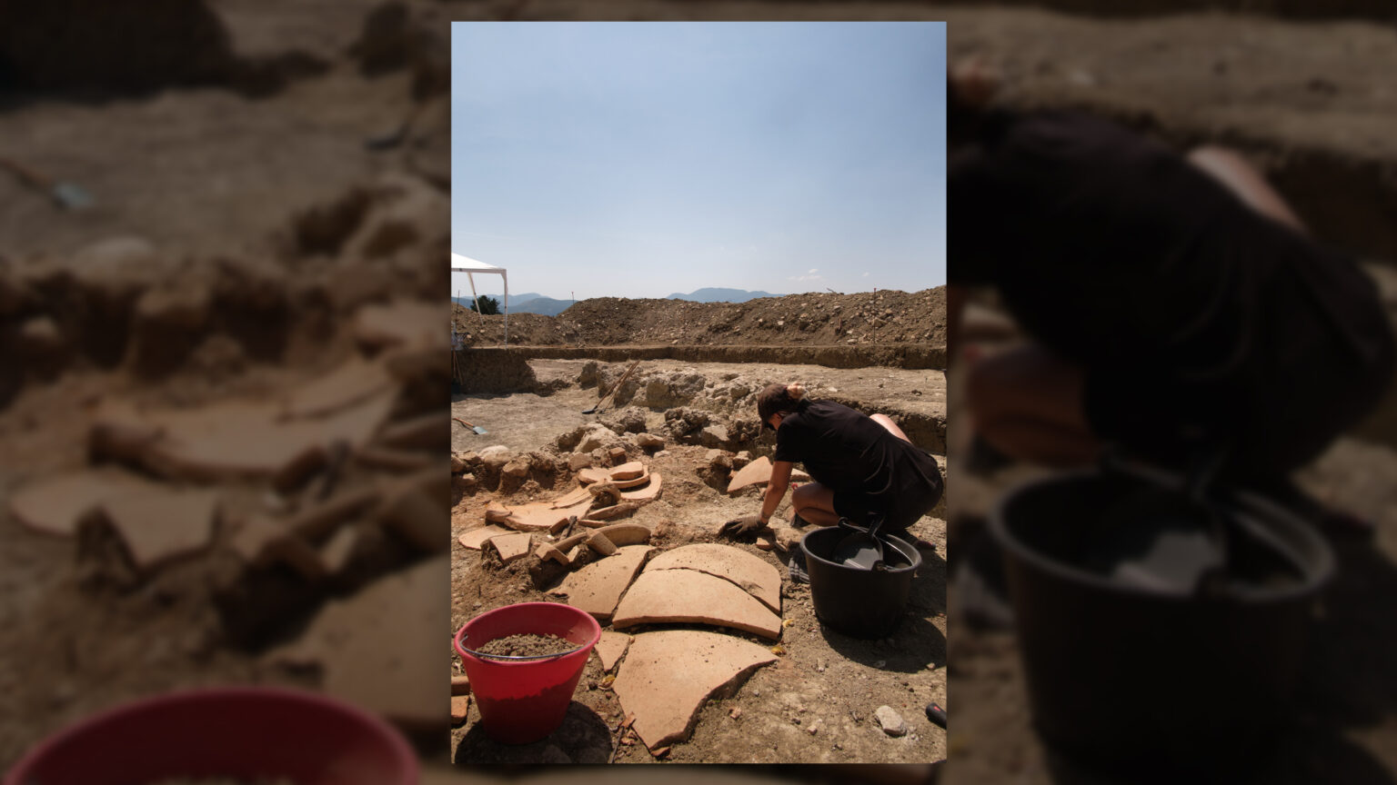 A person crouches down to excavate pottery in a dirt trench.