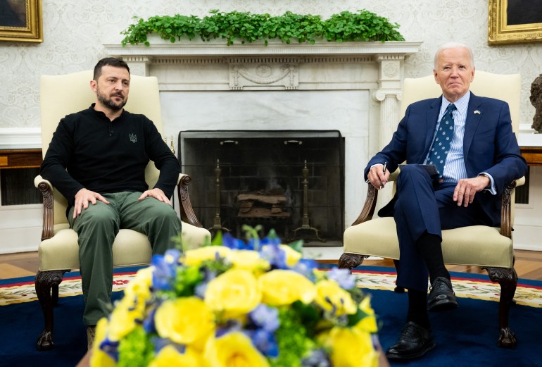 Volodymyr Zelenskyy and Joe Biden in the Oval Office. The are sitting in front of a fire place. There is a large display of blue and yellow flowers on a table in front of them.