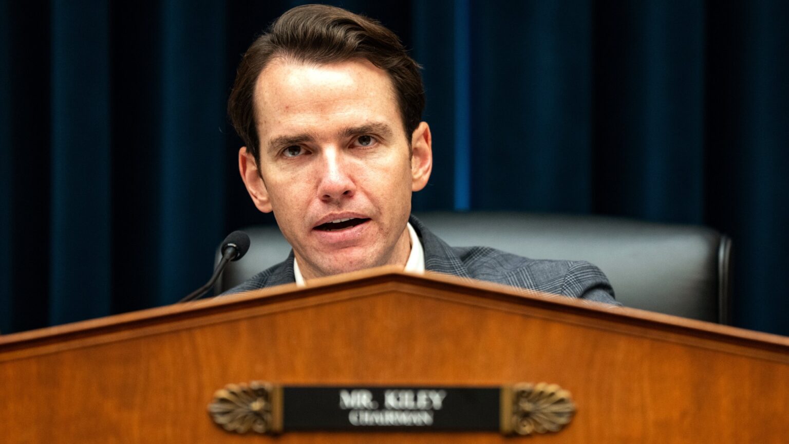 a man in a suit speaks into a microphone in a large hearing room