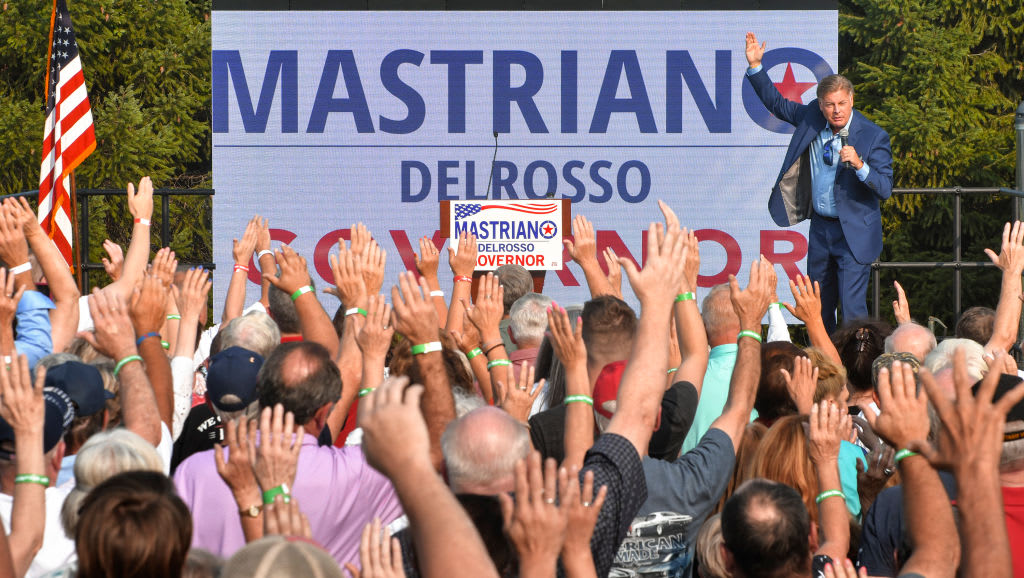 Lance Wallnau campaigns on stage with signs for Doug Mastriano in background.