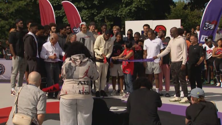 At an outdoor basketball court on a sunny day, photographers snap shots of Vince Carter and Mayor Olivia Chow cutting the ribbon on the refurbished courts, while surrounded by a crowd from the Raptors organization