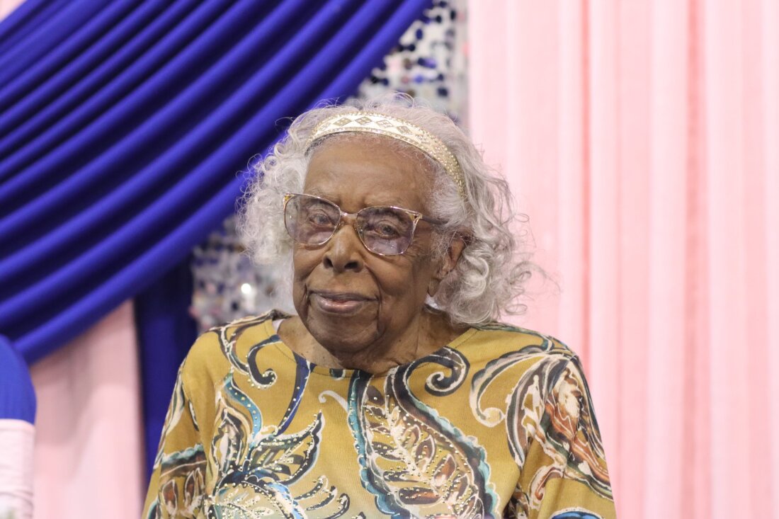 This photo shows Lillie Cunningham. She is wearing glasses and a yellow-brown shirt with a leafy pattern on it. She has gray hair and is wearing a white and gold hair band.