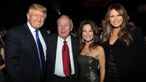 Four smiling people in a row, three with light-toned skin and one with light-medium-toned skin. The two on the left are men and the two on the right are women. The two on the left wear suits while the other two wear dresses.