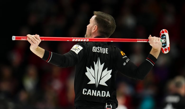 A male curler wearing a black shirt with his name and Canada written at the back of it holds his broom on his back as he looks on.