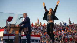 A man joyously jumps in the air on the right as he wears a sportcoat with a T-shirt underneath, behind a man speaking at a lectern.