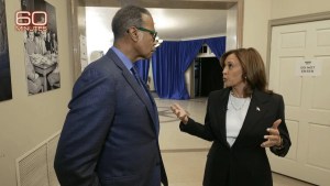 A man and a woman with dark-toned skin stand in a hallway, wearing suits.