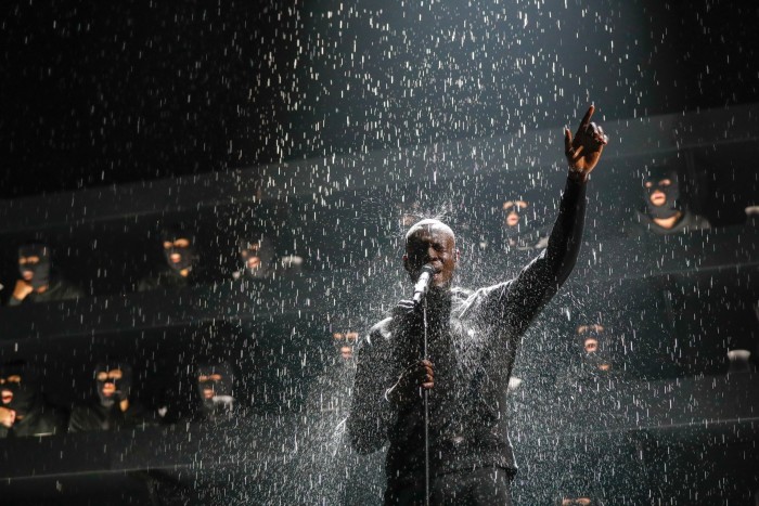 A man standing in front of a microphone under heavy rainfall with one arm outstretched above him