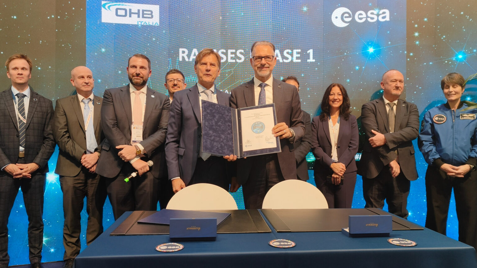 Ten well-dressed people stand behind a table, one of them holding a fancy document