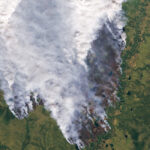 Satellite view of green and brown landscape and snaky river with smoke from forest fires covering most the image.