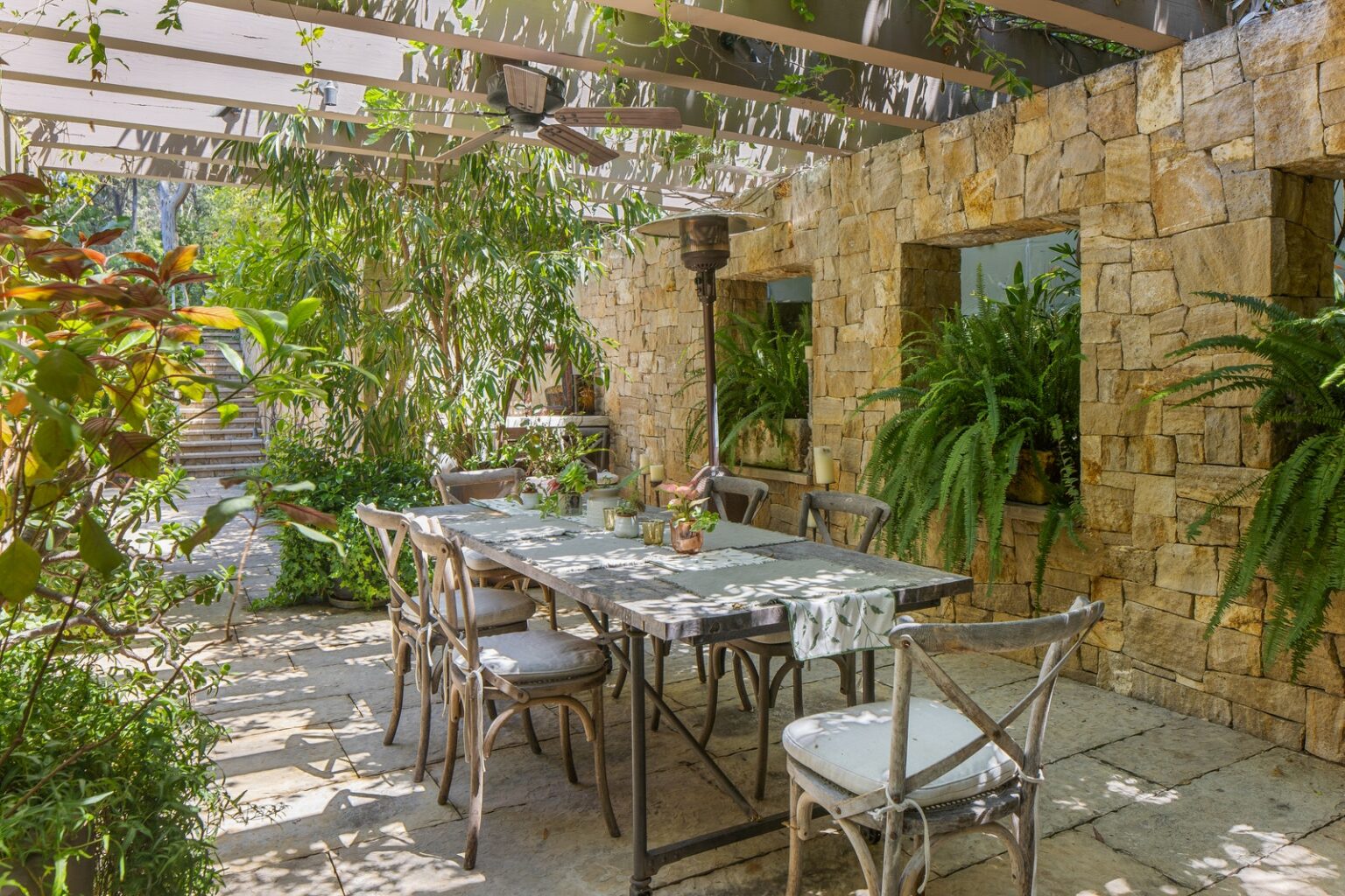 dining area beneath pergola stone facade to right with large ferns growing in window cutouts