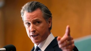 Gavin Newsom, a man with light-toned skin, holds up a finger, gesturing off camera emphatically, in front of a wall with wood paneling