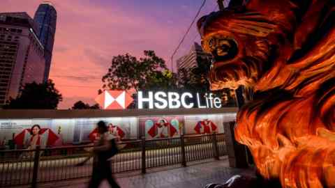 A bronze lion statue is prominently displayed outside the HSBC headquarters in Hong Kong's central financial district. The background shows the HSBC Life sign and modern skyscrapers under a vibrant evening sky.