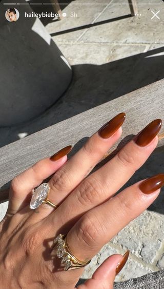 Hailey Bieber shows her autumn leaves manicure in front of a wooden post with two big rings including a 'Mom' ring