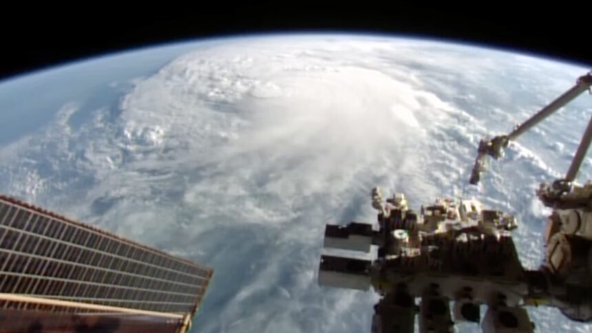 a hurricane and clouds visible on earth. in front is space station infrastructure, a solar panel and a robotic arm