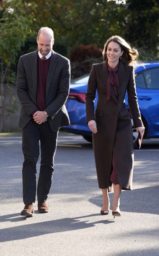 Kate Middleton and Prince William wearing maroon outfits walking through a parking lot