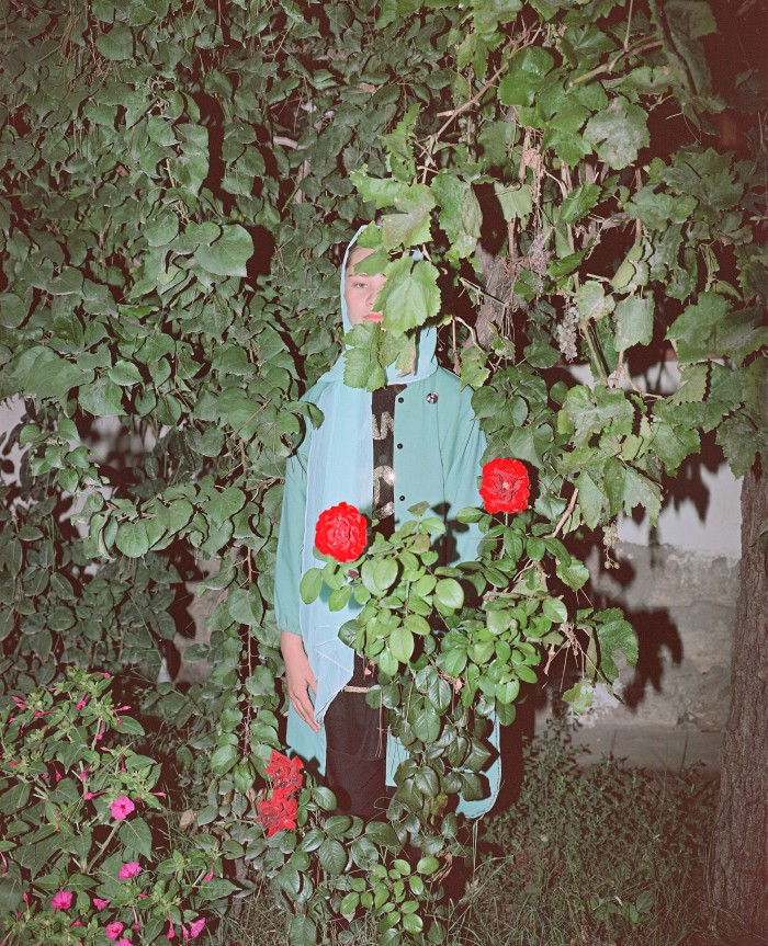 A figure dressed in a blue headscarf and long coat stands amidst thick greenery and vibrant red flowers