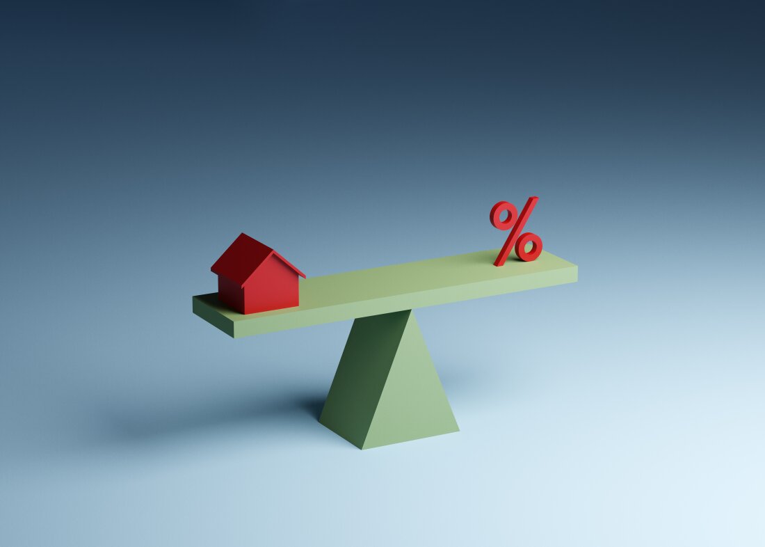 Conceptual illustration of a red toy Monopoly-style house and a red percentage sign balancing on a green seesaw, illustrating the concept of interest rates for a mortgage and refinancing.