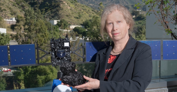 An older white woman in a black blazer standing in front of a model of a spacecraft and holding a model of an outer solar system object.