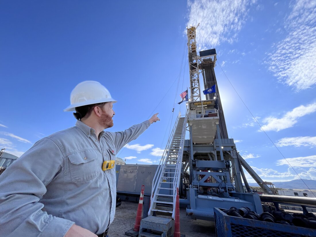 A man wearing a hard hat points upward at a tall drill rig with an American flag hanging from it.