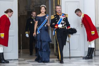 Queen Mary of Denmark wearing a blue evening gown and a pearl and diamond tiara walking arm-in-arm with King Frederik wearing a black military uniform as staff bow to them