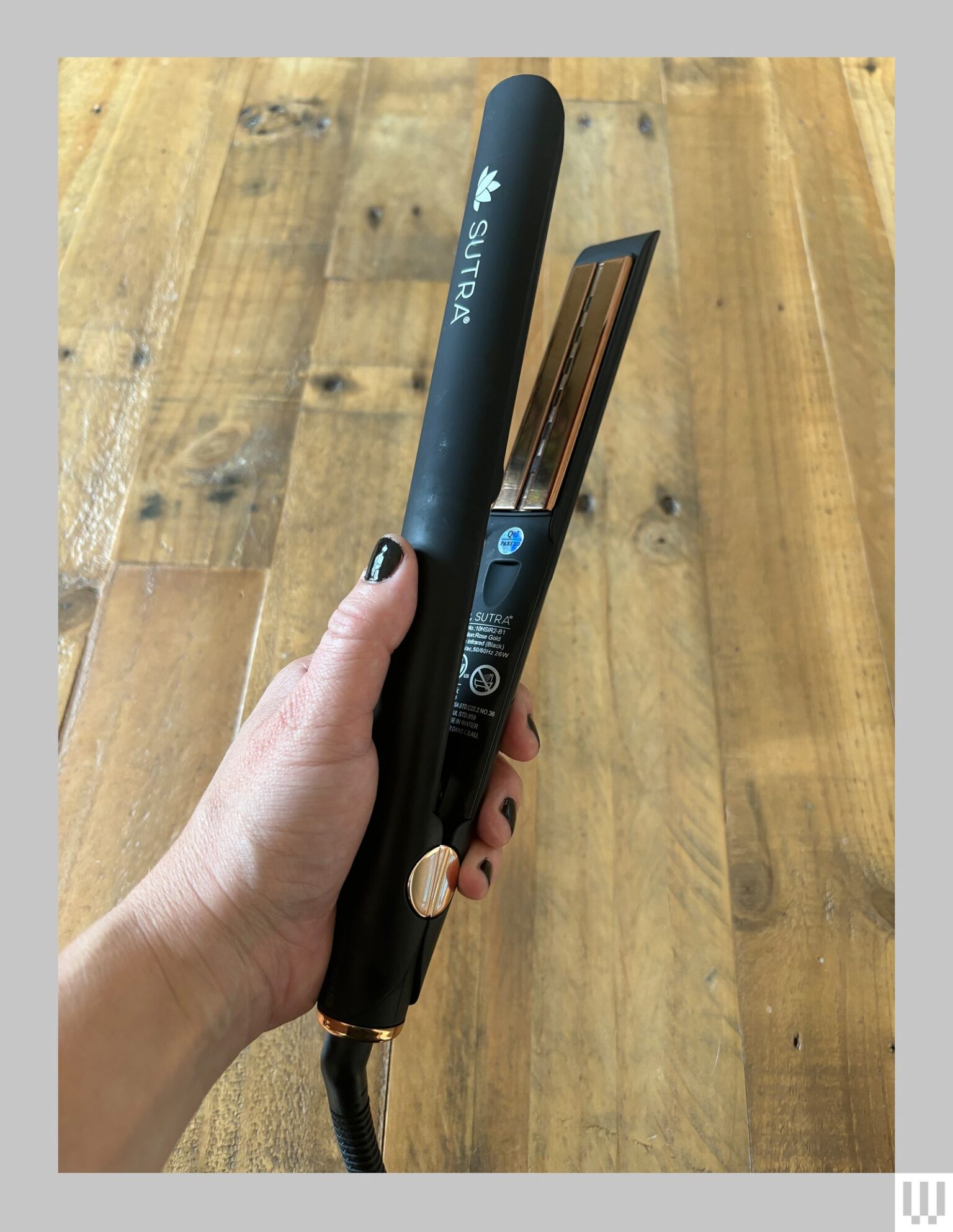 Hand holding a thin black hair straightener showing a gold colored panel on the inside of one of the clamps