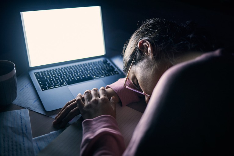 A woman sleeps at a desk, slumped on an illuminated laptop.