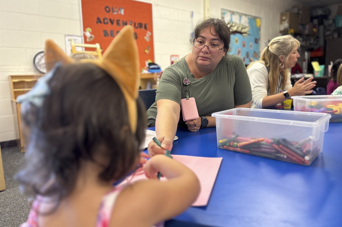 A little girl with her back to the camera sits and draws at a low table the color of blueberries in a classroom. Her preschool teacher leans in with a crayon to help. A plastic bin containing crayons sits between them.