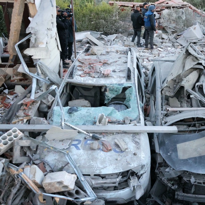 Destroyed cars lie amid rubble at the site of an Israeli air strike. Members of the press can be seen near the wreckage, documenting the aftermath.