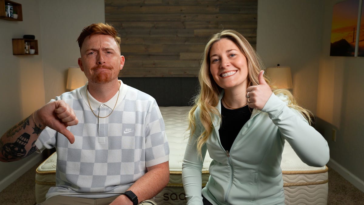 a couple sitting in front of a mattress, the man doing a thumbs-down and frowning, the woman doing a thumbs-up and smiling