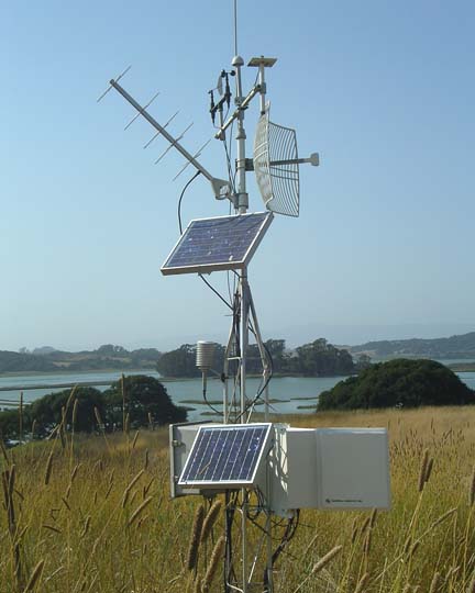 a weather station with several long instruments, flat panels,  and a satellite dish sits in a field near a lake