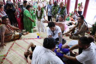 King Charles wearing a white shirt and pants and a lei while watching a group of men tattooing someone's leg on a floor in Samoa