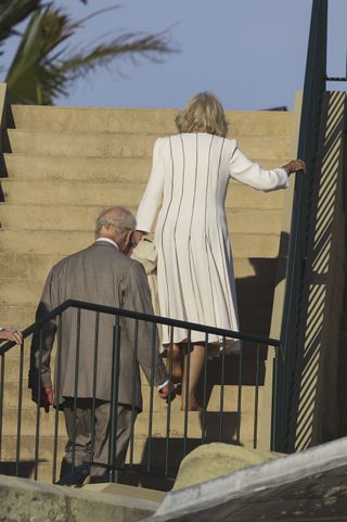 Queen Camilla walking up a staircase outside barefoot followed by King charles wearing a gray suit